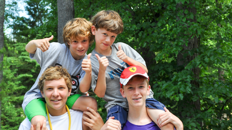 two boys riding on the shoulders of two other boys and giving a thumbs up sign.