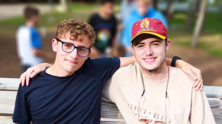 two young men on a bench smiling and holding onto eachothers shoulders.