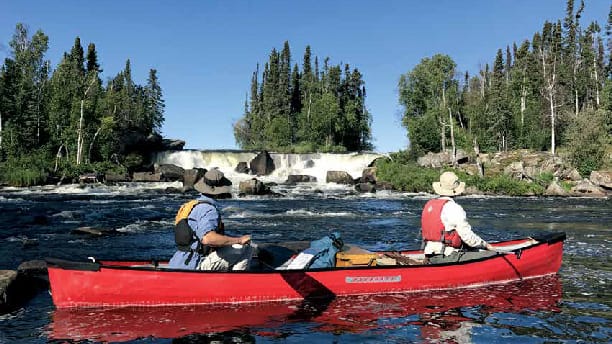 red canoe with two people on it on a river in front of a waterfall.