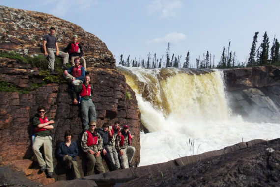 boys standing next to cliff in front of waterfall.