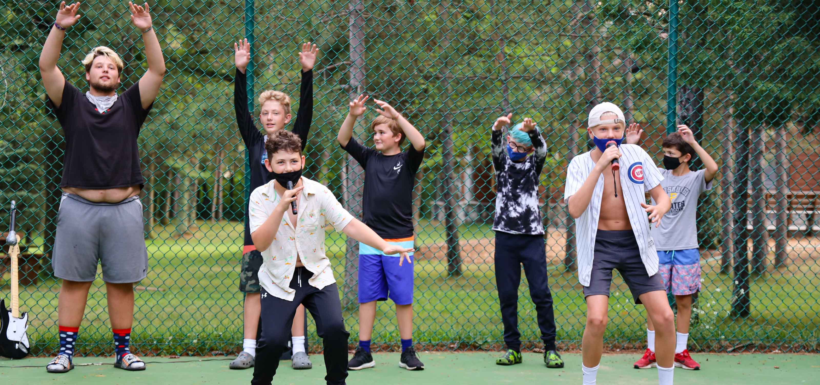 2 boys singing into microphones outside in front of a group of five boys hyping them up.