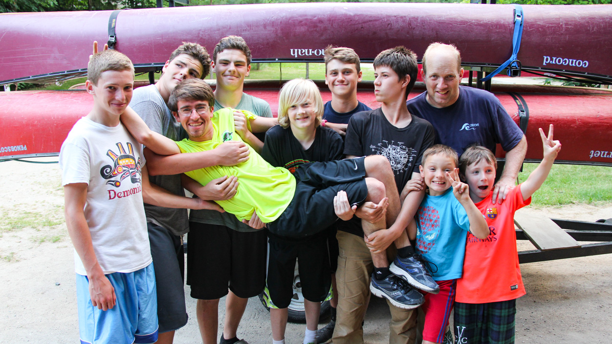 group of boys smiling and holding a camp counselor horizontally in the air.