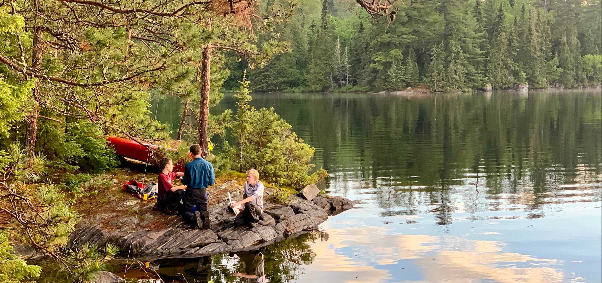 boys with camping gear sitting on a rock next to a lake.