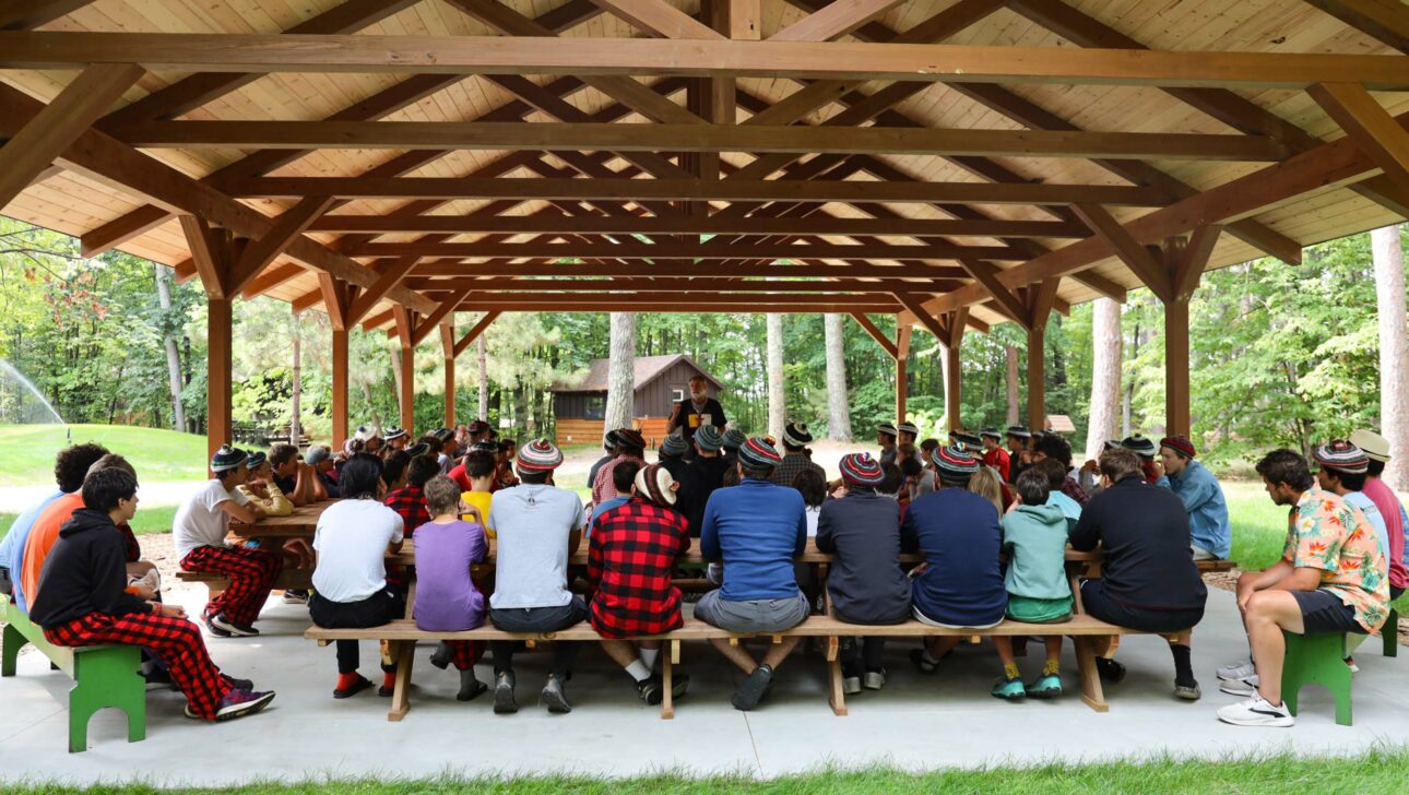 large group of boys sitting at picnic tables underneath pavillion.