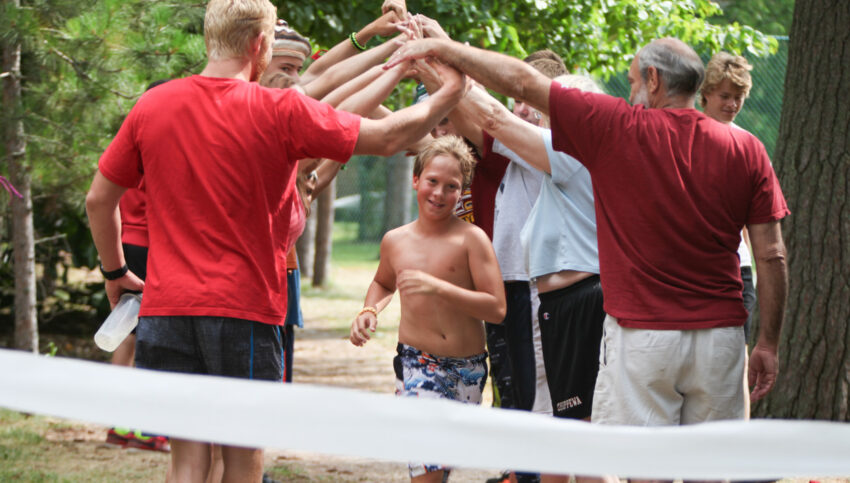 boy running to finish line tape at end of triathalon under arms of camp counselors arched overhead.