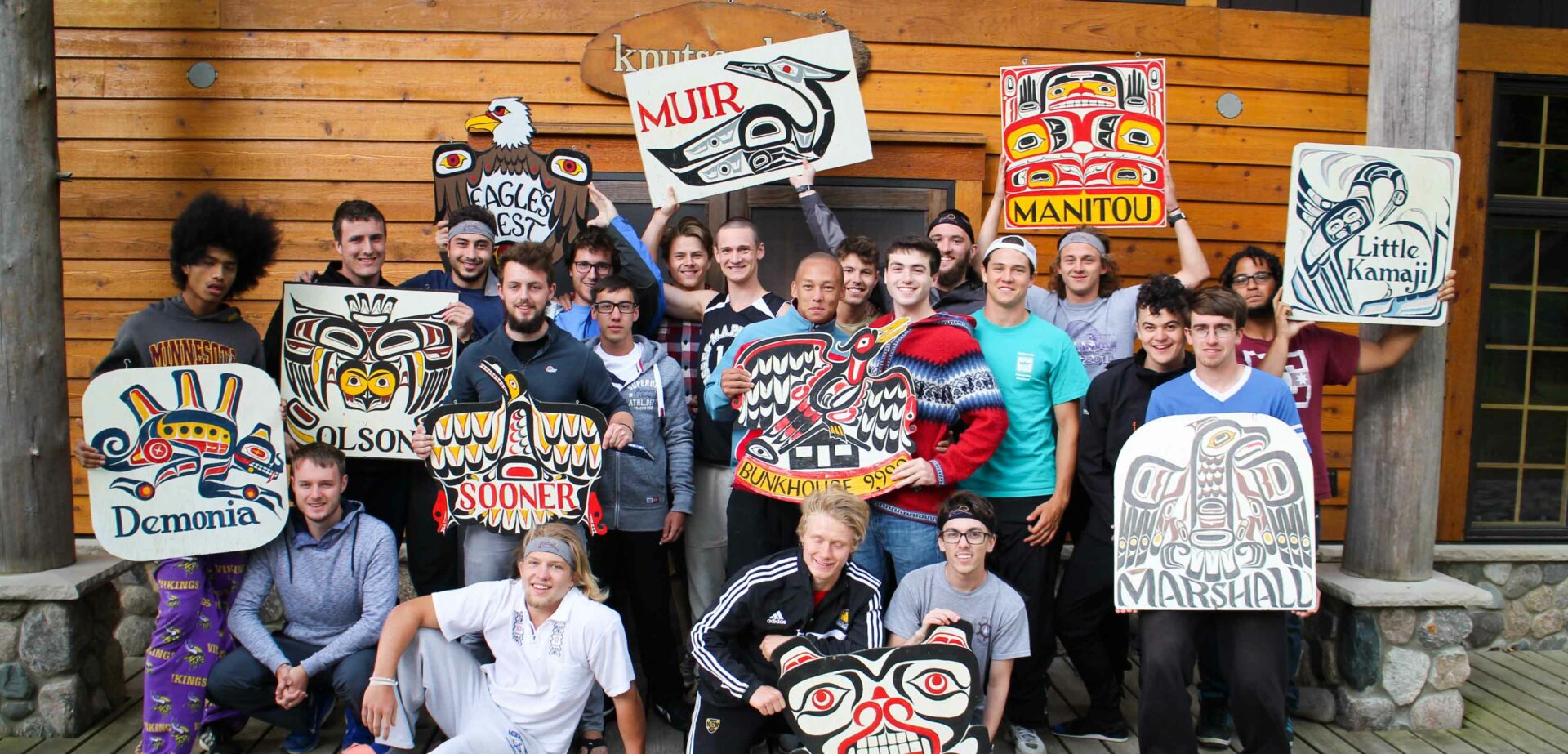 large group of young adult men holding signs in front of a cabin.