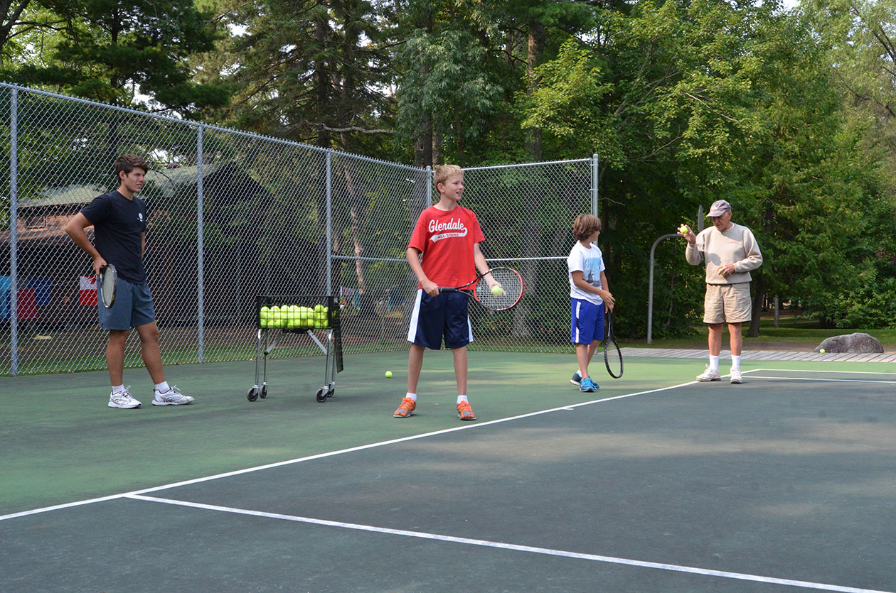 two campers practicing serves on a tennis court.