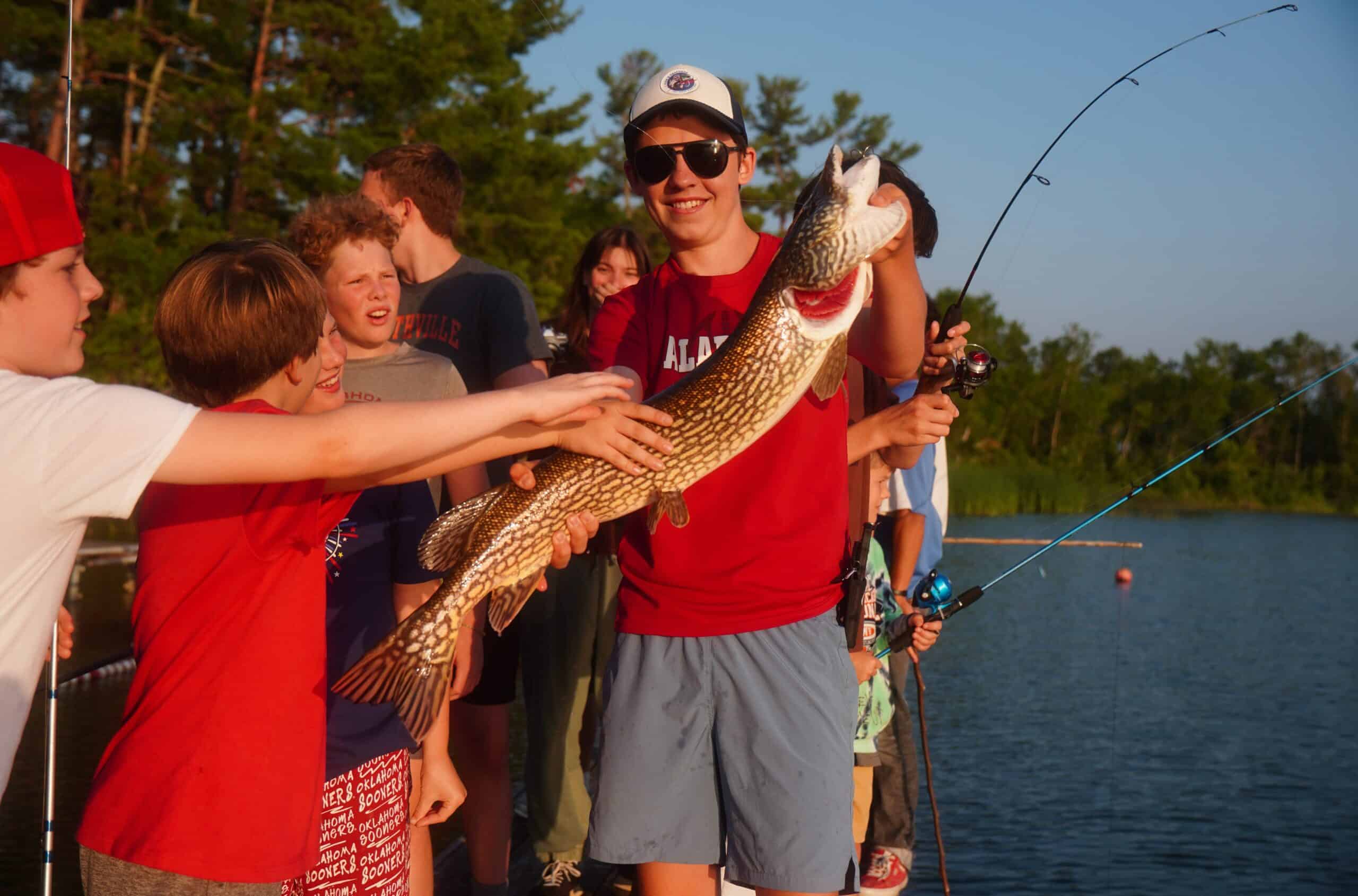 camper smiling and holding large northern pike fish while others watch.