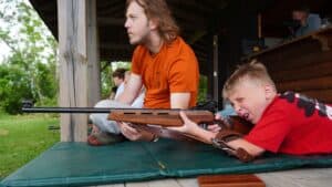 counselor helping camper shoot rifler at summer camp.