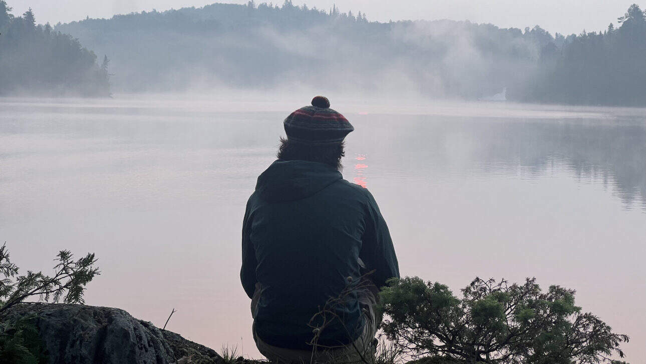 boy with tam sitting on cliff watching sunrise over foggy lake.